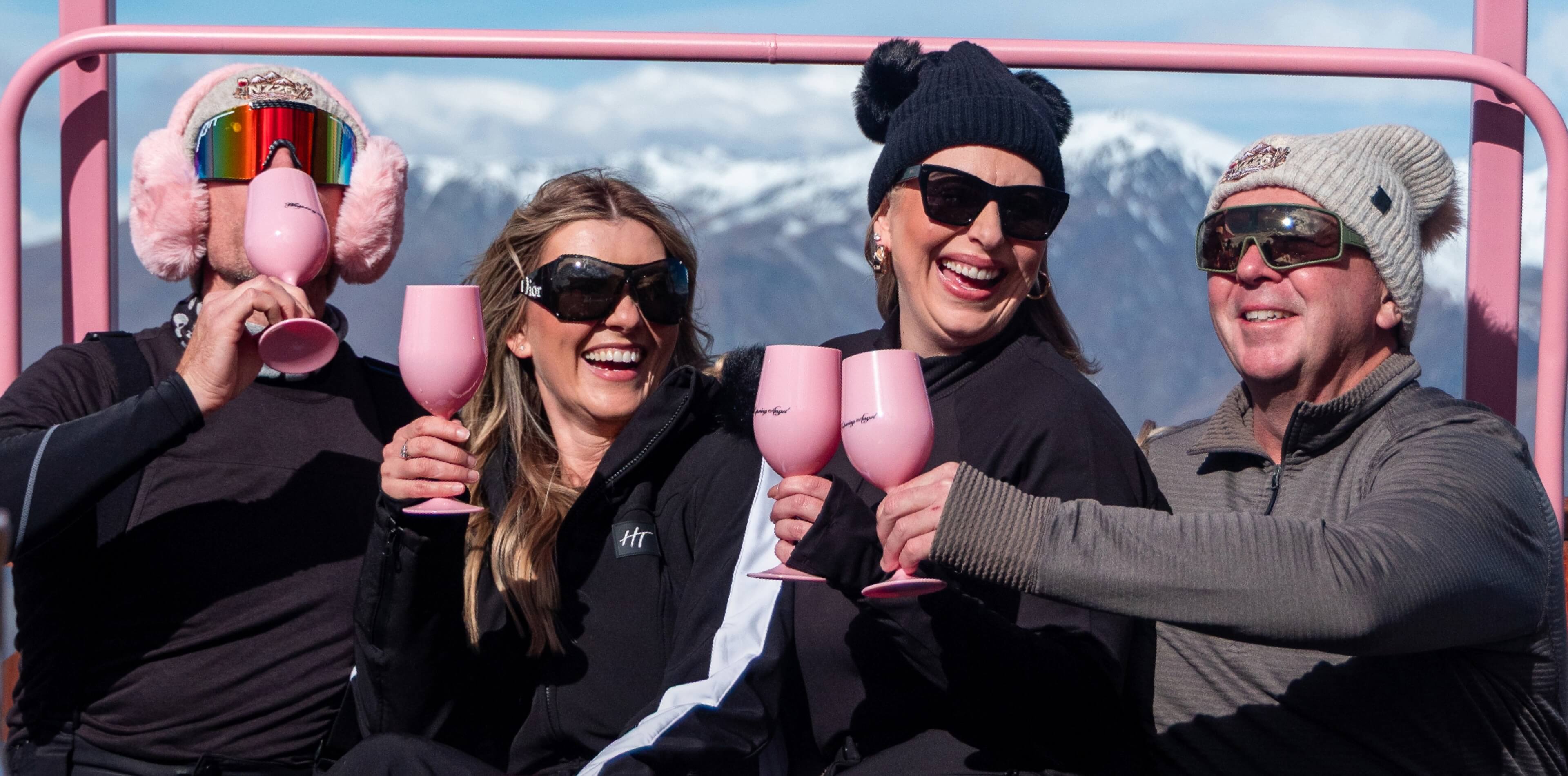 Four adults sitting om a pink bench at Heidi's Hutt on Coronet Peak, smiling and raising pink wine glasses with snowy mountains in the background.